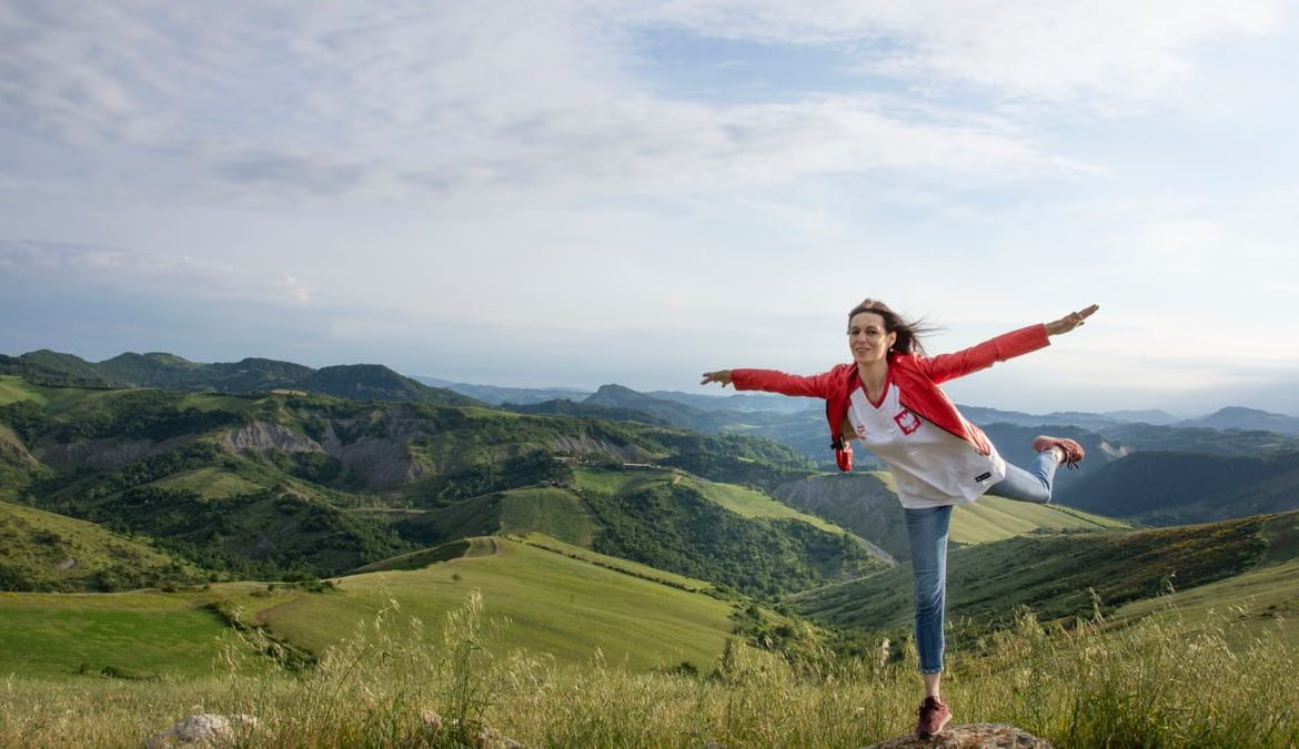 A woman joyfully balances on a rock amidst rolling green hills under a clear blue sky.