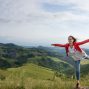 A woman joyfully balances on a rock amidst rolling green hills under a clear blue sky.