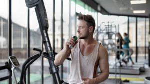 Muscular young sportsman in casual sportswear drinking fresh energy beverage near window while resting after intense workout in modern fitness club